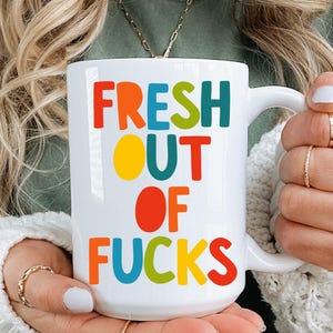 Op de afbeelding: Witte keramische mok met de tekst "FRESH OUT OF FUCKS" in kleurrijke blokletters. De mok heeft een grote handgreep. De mok wordt vastgehouden voor een persoon die een groen shirt en een witte trui draagt.