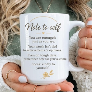 May include: White ceramic mug with the text "Note to self" and inspirational quotes. The mug has a curved handle and a floral design at the bottom. The mug is held by a person wearing a green shirt and a white sweater.