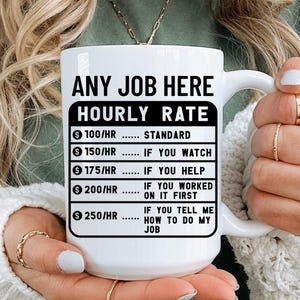 Pode incluir: Caneca de cerâmica branca com o texto "ANY JOB HERE HOURLY RATE" e uma lista de taxas horárias com base no desempenho no trabalho. A caneca é segurada por uma pessoa que usa uma camisa verde e um suéter branco.