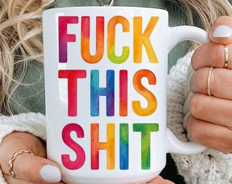 May include: White ceramic mug with the text "FUCK THIS SHIT" in rainbow colours. The mug has a white handle and is held by a person wearing a green top and a white cardigan.