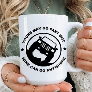 May include: White ceramic mug featuring a black graphic of a 4x4 vehicle and the text "Yours May Go Fast But Mine Can Go Anywhere". The mug is held by a person wearing a green top and a white cardigan.