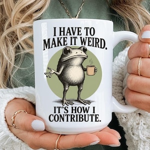 Pode incluir: Uma caneca de cerâmica branca com uma ilustração gráfica de um sapo segurando uma pequena xícara. O texto acima do sapo diz I HAVE TO MAKE IT WEIRD. e o texto abaixo diz IT'S HOW I CONTRIBUTE. O sapo está sobre um círculo verde.