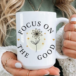 Puede incluir: Taza de cerámica blanca con el texto "FOCUS ON THE GOOD" en negro, que rodea un diseño floral. El diseño incluye una margarita con un centro amarillo y detalles dorados. La taza tiene un asa grande.