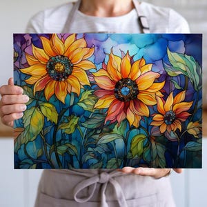May include: A vibrant floral artwork featuring three sunflowers with yellow and orange petals, blue centers, and green leaves. The background has a watercolor effect with shades of blue, purple, and pink. The artwork is held by a person.