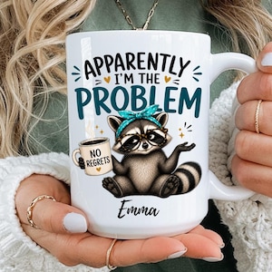 Może przedstawiać: Biały ceramiczny kubek z grafiką szopa pracza w turkusowej opasce na głowie, trzymającego małą filiżankę kawy. Napis brzmi "APPARENTLY I'M THE PROBLEM" w kolorze czarnym i turkusowym, z "NO REGRETS" na filiżance oraz imieniem "Emma" poniżej.