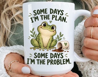 May include: A white ceramic mug featuring a green frog illustration and the text "SOME DAYS I'M THE PLAN. SOME DAYS I'M THE PROBLEM." The design includes a small spilled mug graphic next to the frog, with green leaves and a tiny heart accent.