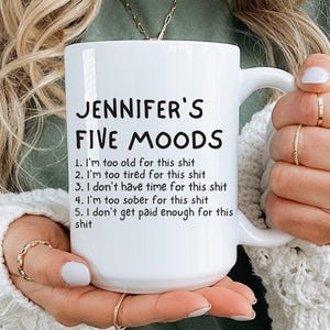 May include: White ceramic mug with the text "JENNIFER'S FIVE MOODS" and a list of five humorous statements. The mug is held by a person wearing a green shirt and a white sweater.