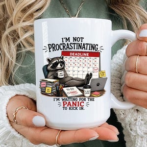 May include: A white ceramic mug featuring a graphic of a raccoon sitting at a desk with a calendar, a laptop, and a stack of papers. The text reads: I'M NOT PROCRASTINATING, I'M WAITING FOR THE PANIC TO KICK IN. The design includes a red deadline banner.
