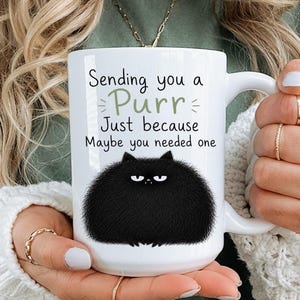 May include: White ceramic mug with a black fluffy cat illustration and the text "Sending you a Purr Just because Maybe you needed one." The mug has a curved handle and is held by a person.
