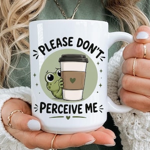 Peut inclure: Mug en céramique blanche avec l'inscription "PLEASE DON'T PERCEIVE ME" et une grenouille de dessin animé cachée derrière une tasse à café. La tasse a une grande anse et est tenue par une personne.