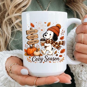 May include: A white ceramic mug featuring a cartoon dog wearing an orange knit hat and scarf, holding a coffee cup. The design includes orange pumpkins, autumn leaves, and wooden signs reading Coffee, Cozy, and Good Vibes with Cozy Season text.
