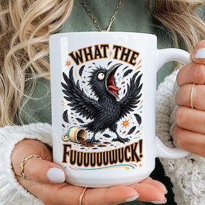 May include: A white ceramic mug featuring a graphic of a black crow with its wings spread wide, a spilled cup, and the text "WHAT THE FUUUUUWUCK!" in bold brown lettering, accented with small colorful stars and decorative flourishes.