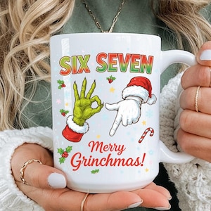 Op de afbeelding: Witte keramische mok met de tekst "SIX SEVEN" in rood en groen. De mok heeft Grinch-thema-illustraties, waaronder een groene hand en een hand met een kerstmuts. De woorden "Merry Grinchmas!" staan ook op de mok.