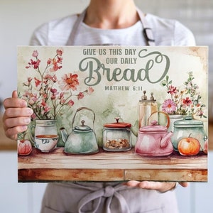 May include: A watercolour painting of a kitchen scene with a quote from the Bible, "Give us this day our daily bread, Matthew 6:11". The painting features a wooden table with a teapot, a jar, and a jug, all in pastel colours. There are also flowers and apples on the table.