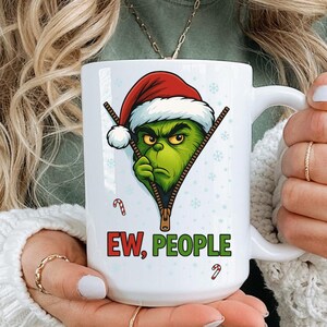 Puede incluir: Taza de cerámica blanca con un personaje verde de Grinch con gorro de Papá Noel, saliendo de un diseño de cremallera. La taza tiene el texto "EW, PEOPLE" en rojo y verde, con detalles de bastones de caramelo. Un diseño humorístico con temática navideña.