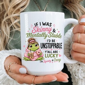 May include: White ceramic mug with the text "IF I WAS Skinny & Mentally Stable I'D BE UNSTOPPABLE Y'ALL ARE LUCKY I'M Squishy & Crazy". The mug features a cartoon frog wearing a pink hoodie and holding a drink.