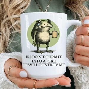 Puede incluir: Taza de cer&aacute;mica blanca con una rana de dibujos animados sosteniendo una taza de caf&eacute;, sobre un c&iacute;rculo verde. La taza tiene el texto "IF I DON'T TURN IT INTO A JOKE IT WILL DESTROY ME" en fuente negra. La taza es sostenida por una persona.