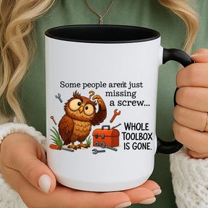 Puede incluir: Taza de cerámica blanca con interior y asa negros. La taza presenta un búho de dibujos animados con herramientas y el texto "Some people aren't just missing a screw... WHOLE TOOLBOX IS GONE."