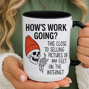 May include: White ceramic mug with a black interior and handle. The mug features the text "HOW'S WORK GOING?" and "THIS CLOSE TO SELLING PICTURES OF MY FEET ON THE INTERNET" along with a skull wearing an orange beanie.