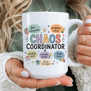 May include: A white ceramic mug featuring the text "CHAOS COORDINATOR" in large, colorful letters. Surrounding the text are smaller speech bubbles with the phrases "fire extinguisher," "problem solver," "schedule saver," "miracle worker," "team hero," and "stress manager," along with small icons like a fire extinguisher, lightbulb, calendar, wand, coffee cup, and people.