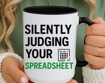 Könnte beinhalten: Weißer Keramikbecher mit schwarzem Innenraum und Henkel. Der Becher zeigt den Text "SILENTLY JUDGING YOUR SPREADSHEET" in Schwarz und Grün, zusammen mit einem Taschenrechner-Symbol. Der Becher wird von einer Person gehalten, die ein grünes T-Shirt und einen weißen Pullover trägt.