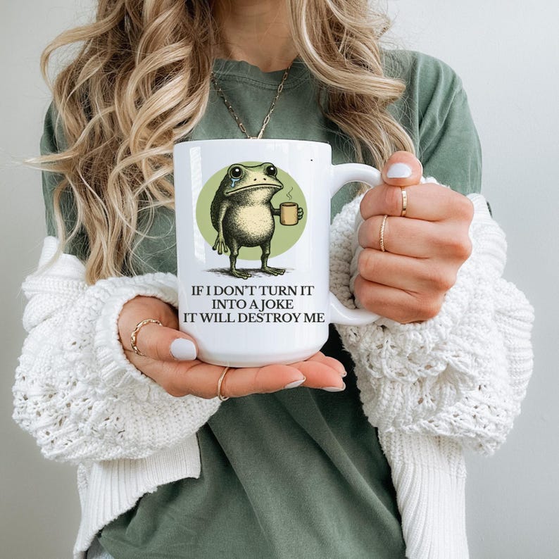 Puede incluir: Taza de cer&aacute;mica blanca con una rana de dibujos animados sosteniendo una taza de caf&eacute;, sobre un c&iacute;rculo verde. La taza tiene el texto "IF I DON'T TURN IT INTO A JOKE IT WILL DESTROY ME" en fuente negra. La taza es sostenida por una persona.