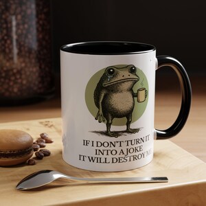 Puede incluir: Taza de cer&aacute;mica blanca con interior y asa negros. La taza presenta una rana de dibujos animados sosteniendo una taza peque&ntilde;a, con el texto "IF I DON'T TURN IT INTO A JOKE IT WILL DESTROY ME".