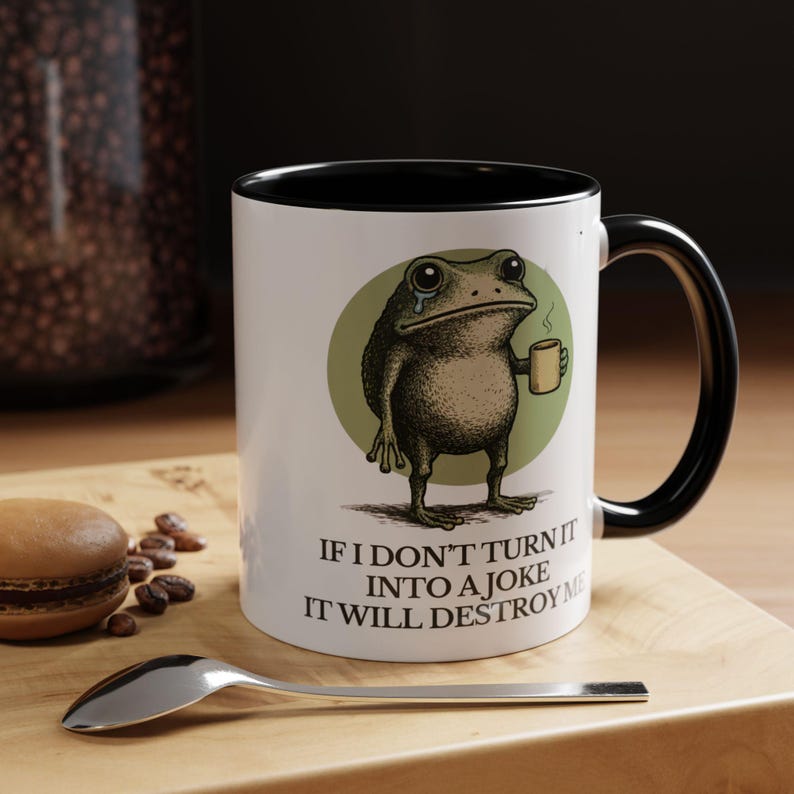 Puede incluir: Taza de cer&aacute;mica blanca con interior y asa negros. La taza presenta una rana de dibujos animados sosteniendo una taza peque&ntilde;a, con el texto "IF I DON'T TURN IT INTO A JOKE IT WILL DESTROY ME".