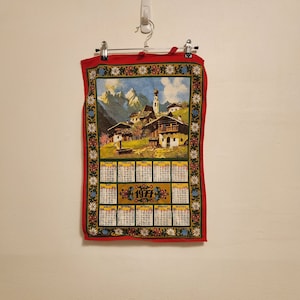 Può includere: Asciugamano da cucina rosso con un bordo floreale e un design di calendario vintage che rappresenta una scena di un villaggio di montagna e l'anno 1971.