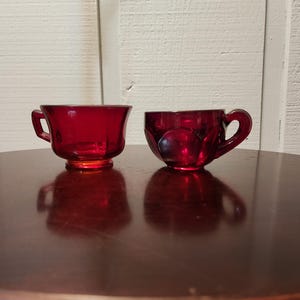 May include: Two red glass cups, one with a handle and one without, on a dark brown wooden surface.