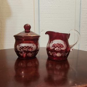 May include: A ruby red glass sugar bowl with a lid and a matching creamer. Both pieces feature a white etched design. The sugar bowl is round with a lid and a small knob. The creamer has a handle and a spout.