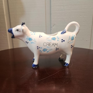 Peut inclure: Une crémière en céramique blanche en forme de vache avec des détails bleus. La crémière en forme de vache présente des motifs floraux bleus et le mot "CREAM". La crémière repose sur quatre pattes et possède une anse.