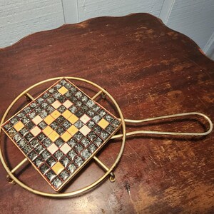 Peut inclure: Un dessous de plat carré composé d'une mosaïque de petites tuiles gris foncé et jaune clair disposées en motif géométrique, inséré dans un cadre circulaire en métal doré avec une longue poignée latérale pour faciliter la manipulation.