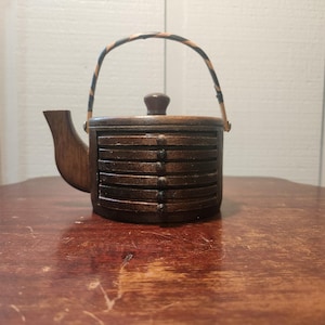 Op de afbeelding: Een donkerbruine theepotvormige houder van hout met een stapel van zes ronde houten onderzetters. De houder is voorzien van een gebogen tuit, een kleine knop op het deksel en een dun, gebogen handvat met een zwart-bruin gestreept patroon.