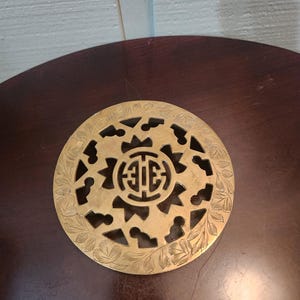 Może przedstawiać: Okrągły, mosiężny podkładka pod garnek z misternym, wyciętym wzorem. Podkładka ma centralny symbol otoczony kwiatowymi i geometrycznymi wzorami. Metal ma ciepły, złoty odcień, a wzór jest szczegółowy i ozdobny.