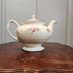May include: Cream-colored porcelain teapot with a lid and handle. The teapot features floral designs in pink, purple, and green. A gold band encircles the base and the lid. The teapot is placed on a dark wooden surface.