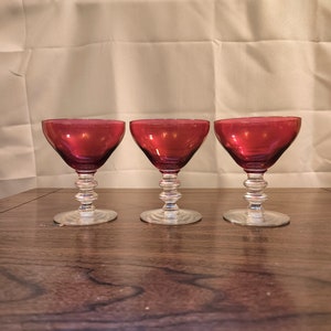 Könnte beinhalten: Drei klare, gestielte Trinkgläser mit einem rubinroten Farbton. Die Gläser haben eine abgerundete Schale und einen klaren, strukturierten Stiel.