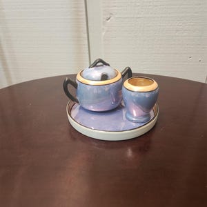 Puede incluir: Un juego de azúcar y crema de porcelana azul y dorado con una tapa en el azucarero. El juego está sobre un platillo blanco con un borde negro.