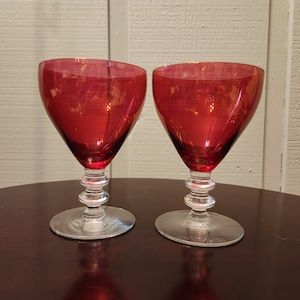 Pode incluir: Duas taças de vinho tinto com hastes e bases de vidro transparente. As taças têm uma forma de tigela arredondada.