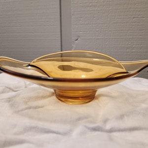 May include: A small, amber-colored glass dish with a unique, wave-like design. The dish has a wide, shallow bowl and a small, rounded base.