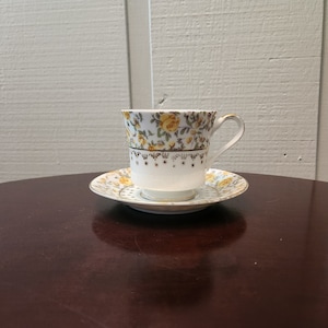 Puede incluir: Una taza de té y un platillo blancos con un patrón floral con rosas amarillas y hojas verdes. La taza y el platillo tienen un borde dorado.