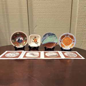 May include: Five small decorative plates with various designs, including a blue and white floral pattern, a red and white floral pattern, a blue and white geometric pattern, and a red and white geometric pattern. The plates are displayed on a brown wooden table.