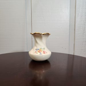 May include: A small, white ceramic vase with a gold rim and floral pattern. The vase has a scalloped top and a rounded body.