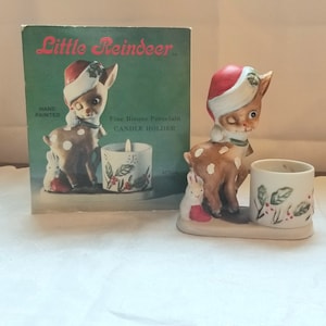 Op de afbeelding: Een handgeschilderde bisque porseleinen kaarsenhouder met een rendier dat een kerstmanmuts draagt en een wit konijn. De kaarsenhouder is gelabeld "Little Reindeer Fine Bisque Porcelain Candle Holder".