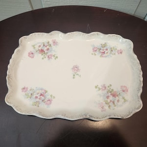May include: An off-white ceramic serving tray, rectangular with a scalloped edge. It features floral patterns in pink and blue, with a gold trim. The tray is ideal for serving food.