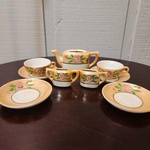 May include: A vintage porcelain tea set with a floral design. The set includes a teapot, two cups, two saucers, and two creamer cups. The tea set is painted with pink and yellow flowers on a cream background.