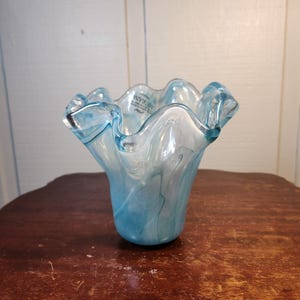 Puede incluir: Un jarrón de cristal transparente con un borde ondulado y un diseño de remolino azul y blanco. El jarrón está sobre una superficie de madera oscura. El jarrón tiene una forma única y es probablemente decorativo.