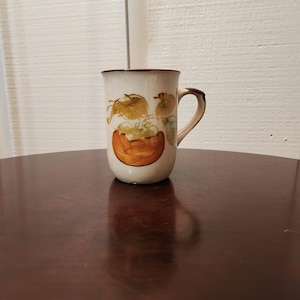 Pode incluir: Uma caneca de cerâmica branca com uma borda e uma alça marrons. A caneca tem um desenho pintado de uma fruta marrom e laranja com folhas verdes.