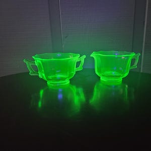 Puede incluir: Un juego de dos azucareros vintage de vidrio verde con asas. Los cuencos están hechos de vidrio de uranio y brillan bajo una luz negra.