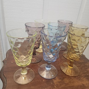 May include: A collection of seven multi-colored glass goblets featuring a diamond-patterned texture. The set includes individual glasses in shades of yellow, purple, light blue, and amber, each supported by a clear glass stem and a circular base.